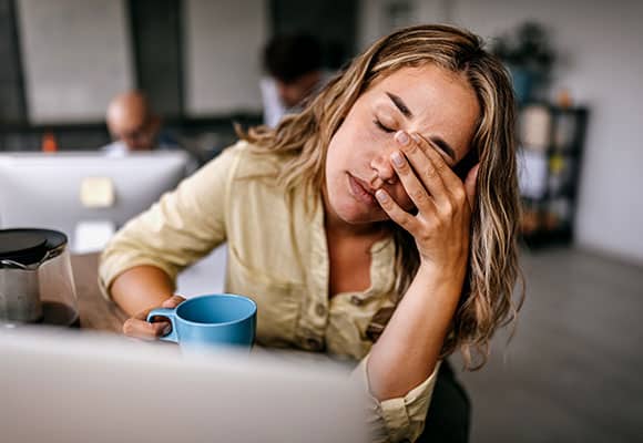 Eine Frau sitzt am Schreibtisch mit einem blauen Becher in der Hand, die Augen geschlossen, eine Hand vor dem Gesicht, müde oder gestresst - vielleicht braucht sie Schüßler Salze, um ihren Mineralstoffhaushalt im Büro zu unterstützen.