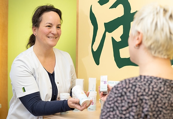 Eine lächelnde Frau in einem weißen Kittel reicht einer anderen Frau über einen Tresen hinweg homöopathische Produkte. Im Hintergrund sind Regale mit weiteren Produkten und eine gelbe Wand mit grüner Schrift zu sehen.