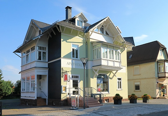 In einem zweistöckigen, hellgrünen Gebäude mit weißer Fassade befindet sich eine Apotheke, die durch das APOTHEKE-Schild und das rote Apothekensymbol neben dem Eingang gekennzeichnet ist. Große Fenster, ein steiles Dach und eine kleine Rampe laden die Besucher zu "Apotheke verstehen" ein.