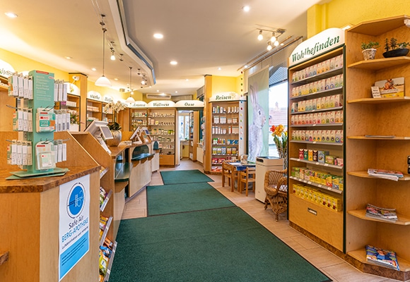Helles, organisiertes Apothekeninterieur mit Holzregalen und grünen Teppichen. In den Regalen stehen Gesundheitsprodukte und Zeitschriften. Große Fenster füllen den Raum mit natürlichem Licht, während deutsche Schilder jede Abteilung beschriften und so für eine einladende, effiziente Atmosphäre sorgen.