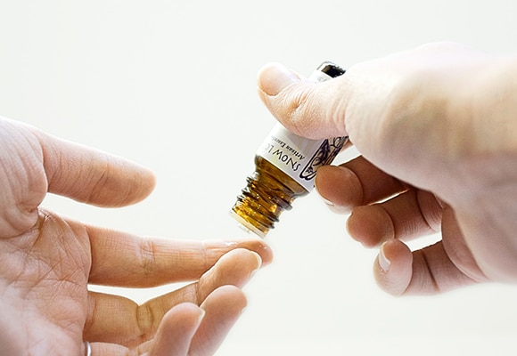 Eine Person gießt eine Flüssigkeit aus einer kleinen braunen Glasflasche auf ihre Fingerspitzen und bereitet sich darauf vor, sie im Rahmen einer Aromatherapie-Routine auf ihre Haut aufzutragen. Der Hintergrund ist schlicht und weiß.
