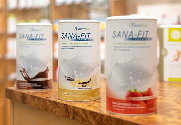 Drei Dosen Bodymed SANA-FIT Premium Mahlzeitenersatzpulver in den Geschmacksrichtungen Schokolade, Vanille und Erdbeere stehen auf einer Holztheke, die das Bodymed Ernährungskonzept präsentiert, wobei die Regale im Hintergrund verschwimmen.