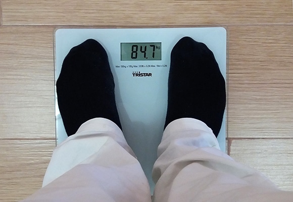 Eine Person, die schwarze Socken und eine helle Hose trägt, steht auf einer digitalen Waage, die auf einem Holzboden ein Gewicht von 84,7 Kilogramm anzeigt, und verfolgt möglicherweise ihre Fortschritte mit dem Bodymed Ernährungskonzept.