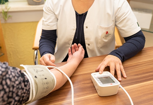 Eine Apothekerin kontrolliert den Blutdruck mit einem elektronischen Messgerät, während der Arm des Patienten auf einem Holztisch liegt.