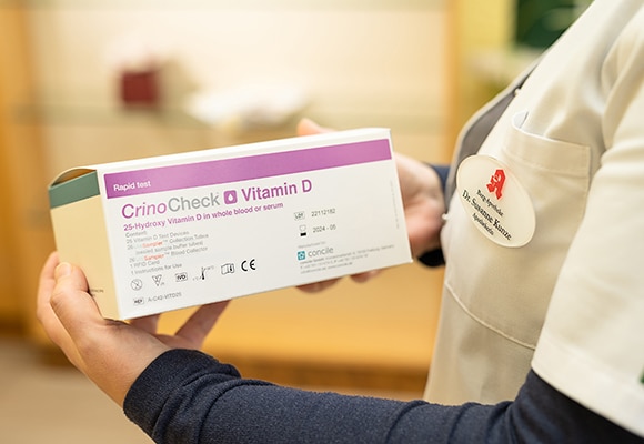 Ein Mitarbeiter der Apotheke hält eine Schachtel mit der Aufschrift CrinoCheck Vitamin-D-Schnelltest für Messungen und Kontrollen von 25-Hydroxy-Vitamin D in Blut oder Serum. Die Person trägt einen weißen Kittel mit Namensschild.