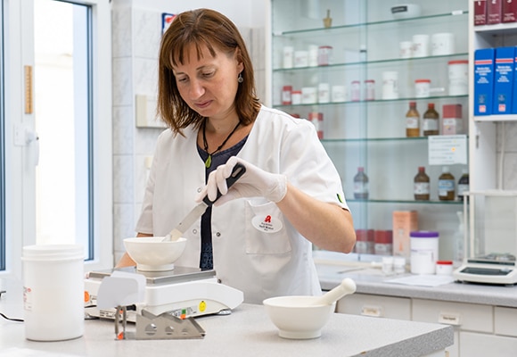 Eine Frau in einem weißen Kittel und mit Handschuhen benutzt einen Spatel, um in einem Mörser in einer Apotheke Substanzen zu mischen oder zuzubereiten, wobei im Hintergrund Regale mit Flaschen und Gläsern zu sehen sind.