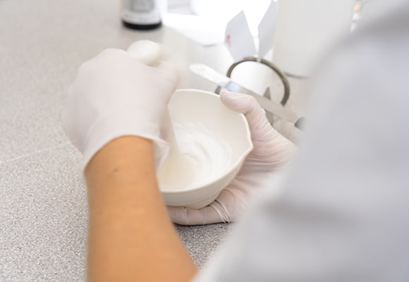 Eine Person mit weißen Handschuhen mischt eine cremig-weiße Substanz in einer kleinen Schüssel auf einer Arbeitsplatte mit einem Spatel in der Rezeptur einer Apotheke.