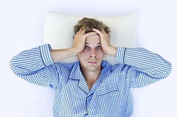 Ein Mann in einem blau gestreiften Schlafanzug liegt im Bett auf einem weißen Kissen, schaut gestresst und mit großen Augen, beide Hände gegen den Kopf gepresst.