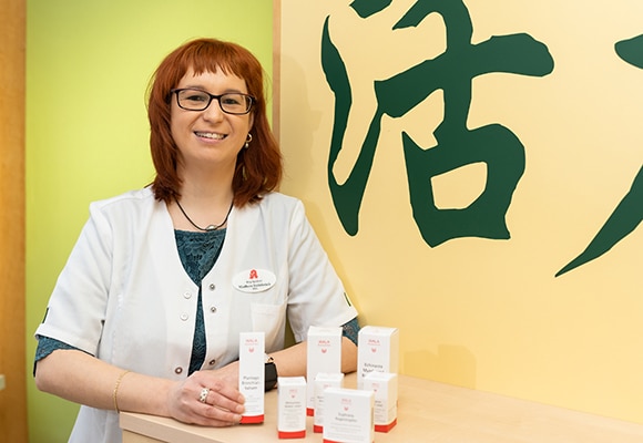 Eine lächelnde Person mit roten Haaren und Brille steht in einem weißen Kittel an einer Auslage für Hautpflegeprodukte. Ein großes, grünes chinesisches Schriftzeichen ist auf der gelben Wand hinter ihnen zu sehen.