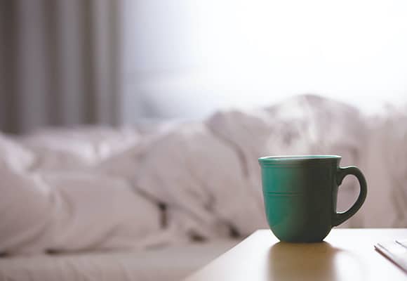 Ein grüner Becher steht auf einem hölzernen Nachttisch vor einem Bett mit zerknitterten weißen Laken, das von natürlichem Licht sanft erhellt wird - eine ruhige Szene, die an die Ruhe erinnert, die oft durch alternative Medizin und Homöopathie gesucht wird.
