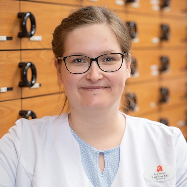Eine Frau, die eine Brille und einen weißen Kittel trägt, lächelt vor einem Hintergrund aus Holzschubladen mit schwarzen Griffen. Sie hat ihr hellbraunes Haar zurückgebunden und trägt ein Namensschild.
