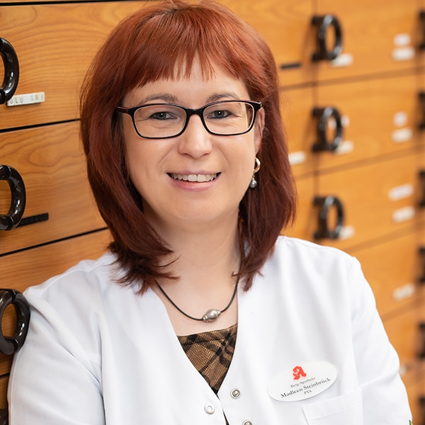 Eine Frau mit schulterlangem rotem Haar und Brille, die einen weißen Kittel und ein Namensschild trägt, steht lächelnd vor hölzernen Schubladen mit schwarzen Griffen in einer Apotheke.