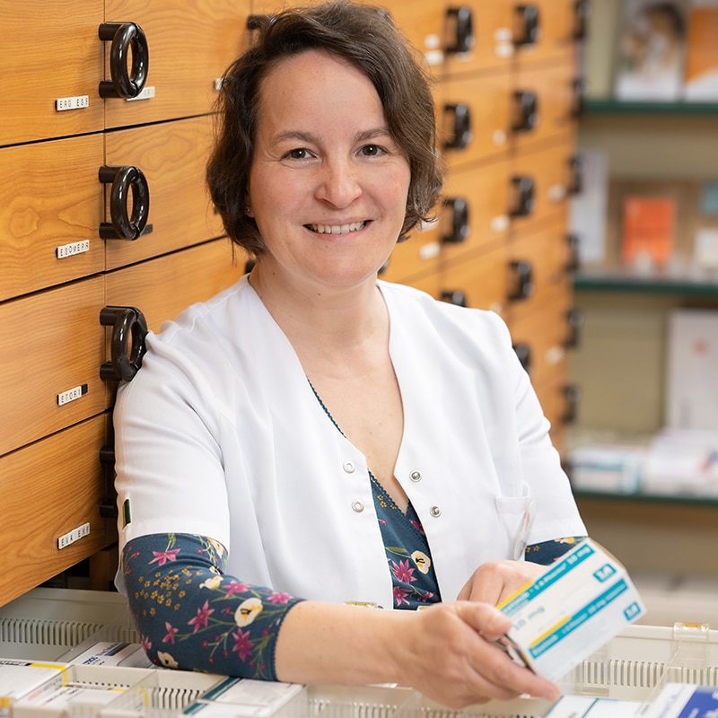 Eine lächelnde Apothekerin im weißen Kittel steht vor hölzernen Schubladen und hält mehrere Kisten mit Medikamenten in der Hand - ein Bild, das das organisierte und professionelle Apothekenkonzept widerspiegelt.