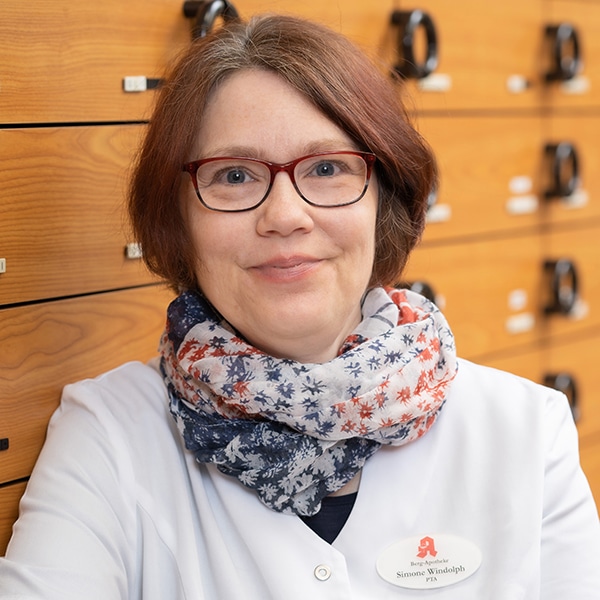Eine Frau mit kurzen braunen Haaren und roter Brille, die einen weißen Laborkittel und einen gemusterten Schal trägt, steht lächelnd vor einer Wand mit Holzschubladen. Sie trägt ein Namensschild an ihrem Kittel.