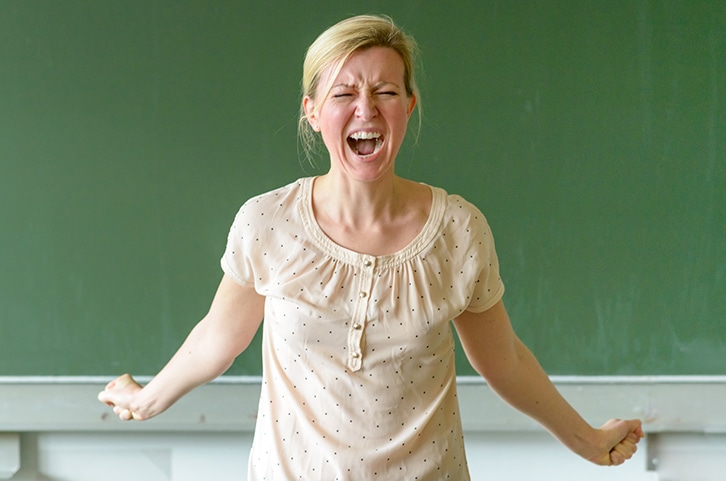 Eine Frau steht vor einer Kreidetafel, ballt die Fäuste und schreit mit einer intensiven Miene, die sichtlich Stress oder innere Unruhe zeigt. Sie trägt eine helle Bluse und hat ihr blondes Haar zurückgebunden.