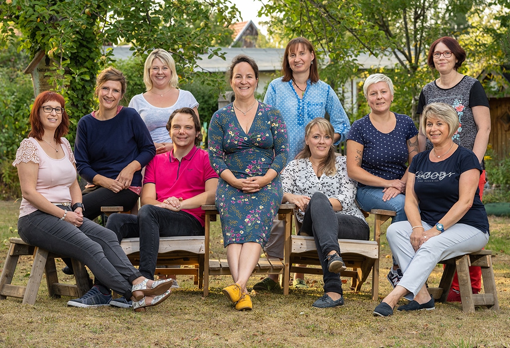Eine Gruppe von zehn Erwachsenen, zumeist Frauen, sitzt und steht im Freien auf Holzbänken zusammen und lächelt in die Kamera in einem Garten mit Bäumen und Grün im Hintergrund.