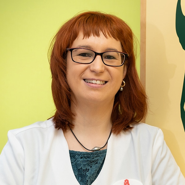 Eine Frau mit schulterlangen roten Haaren und Brille lächelt, während sie einen weißen Kittel über einem blaugrünen Oberteil trägt und vor einem hellgrünen und gelben Hintergrund steht - bereit, Frauen durch Wechseljahre und Hitzewallungen zu begleiten.
