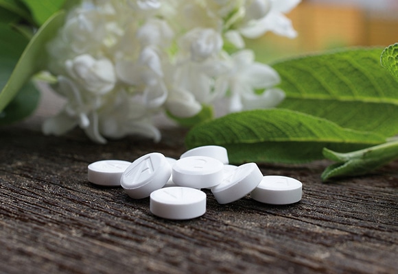 Ein kleiner Stapel weißer, runder Tabletten liegt auf einer hölzernen Fläche, die an Mineralstoffberatung erinnert, mit grünen Blättern und weißen Blumen im Hintergrund.