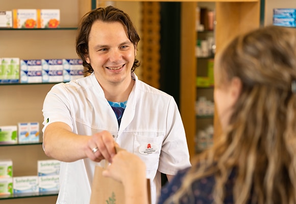 Ein lächelnder Apotheker in einem weißen Kittel reicht einem Kunden über den Tresen hinweg eine Papiertüte, während im Hintergrund Regale mit Medikamenten und Gesundheitsprodukten zu sehen sind.
