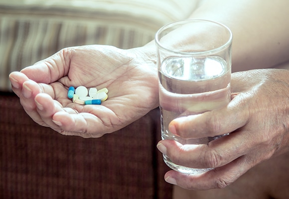 Eine Person hält mehrere Pillen unterschiedlicher Form und Farbe in einer Hand und ein Glas Wasser in der anderen und bereitet sich auf die Einnahme von Medikamenten vor.