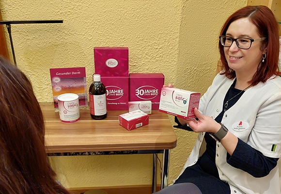 Eine Frau in einem weißen Kittel sitzt an einem Tisch mit rosa Gesundheitsprodukten für die Darmberatung, lächelt und zeigt einer anderen Person eine Schachtel. Die gelbe Wand und die fein säuberlich angeordneten Produktverpackungen schaffen eine einladende Atmosphäre.